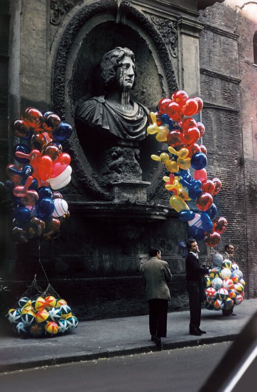 Three men stand on a street near a large stone bust in a wall niche, flanked by colorful clusters of balloons and balls for sale. The scene has an old-world, urban atmosphere with weathered brick and stone.