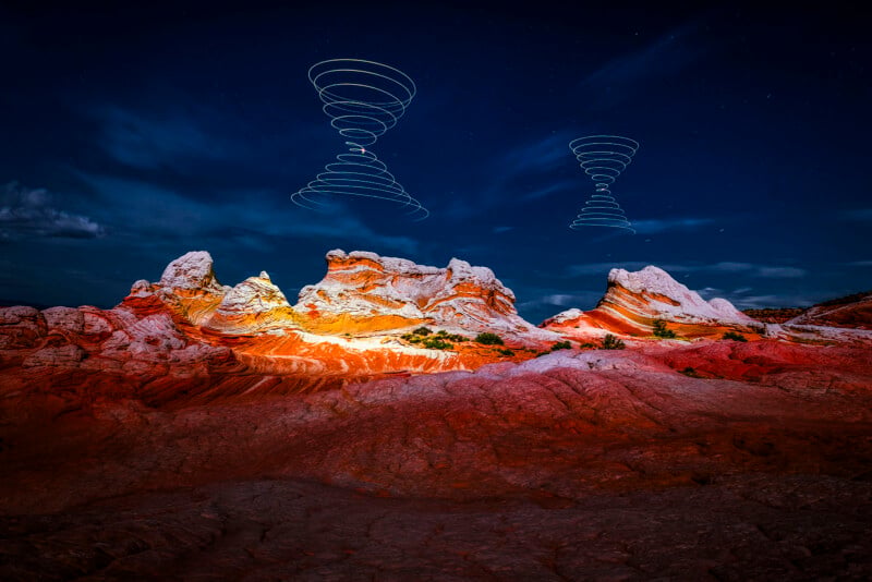 Red and white rock formations under a dark blue night sky, with two spiral light trails floating above the landscape, creating a surreal and otherworldly scene.