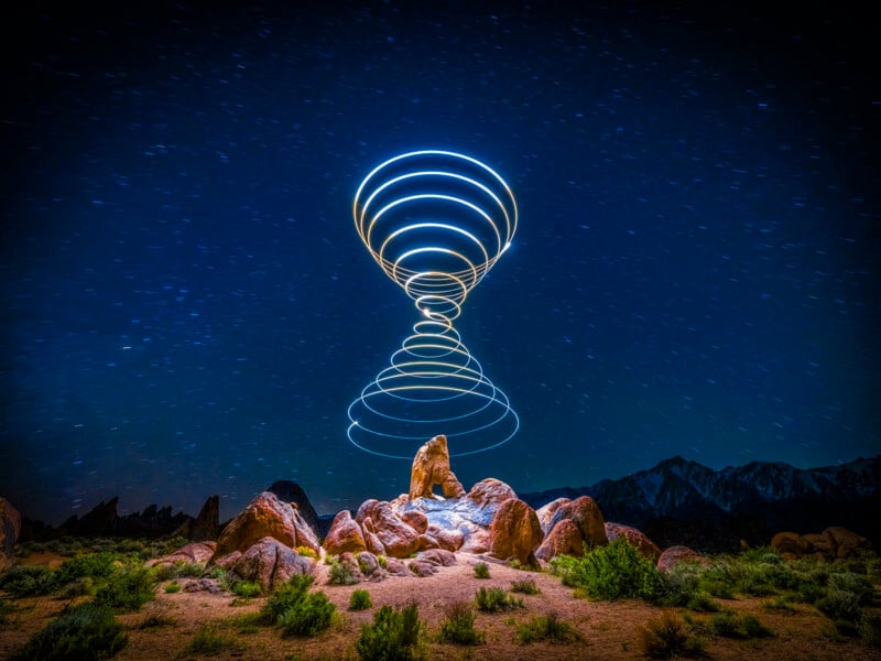 Rocks and shrubs under a night sky, with spiraling rings of light forming an hourglass shape above the landscape; mountains are faintly visible in the background.