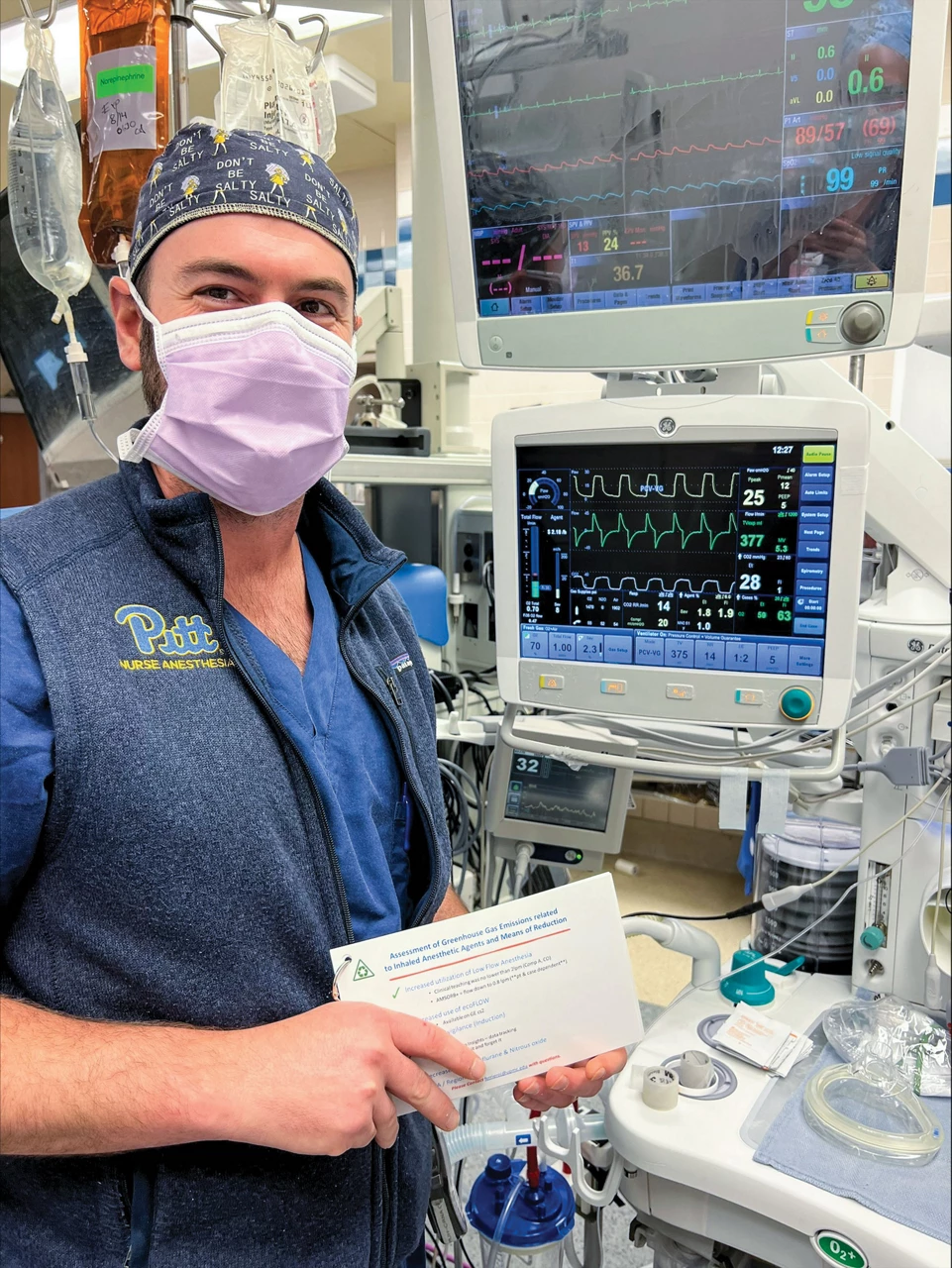 A person wearing a face mask and head covering stands beside a counter containing various pieces of medical equipment, including IV bags and two screens that display data curves.