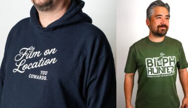 Two men stand side by side. One wears a navy hoodie with “Film on Location You Cowards.” The other wears a green T-shirt reading “BIRD HUNTER Birds I’d Like to Photograph” with a bird graphic above the text.