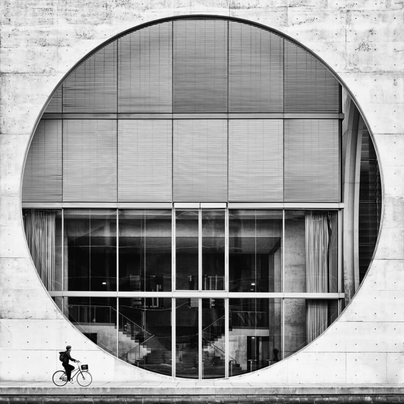 A person rides a bicycle in front of a large modern building with a circular window, showcasing geometric lines and patterns in a black and white composition.