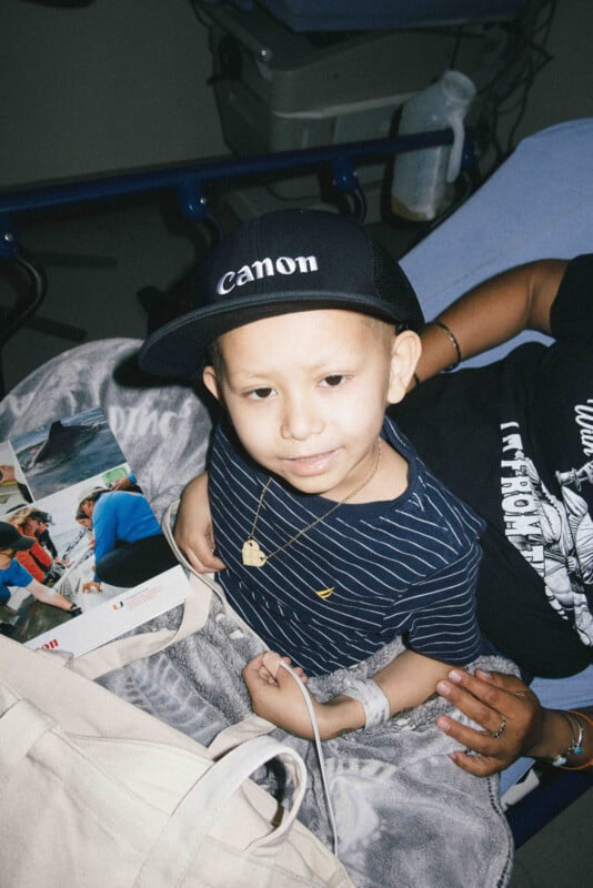A young child wearing a black Canon cap and striped shirt sits on a hospital bed with an IV in his arm, holding a magazine and smiling. An adult, partially visible, gently holds his arm.
