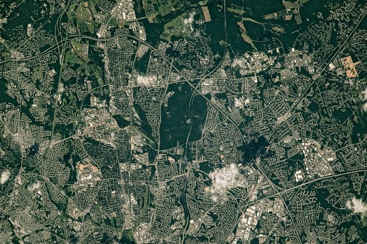A straight-down view of Greenbelt is centered on a square park, with smaller green spaces weaving through surrounding homes, businesses, a college campus, and government buildings.
