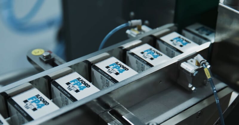 Boxes of Ilford FP4 Plus 125 black-and-white film are lined up on a conveyor belt in a factory setting, ready for packaging or distribution.