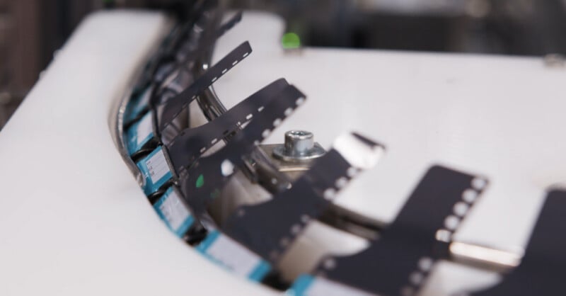 Close-up of a curved conveyor or processing line moving strips of black material with holes along the edges, possibly film or electronic components, over a white surface in an industrial setting.