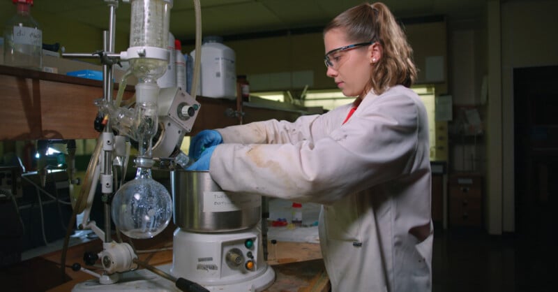 A scientist wearing a white lab coat, safety goggles, and blue gloves works with lab equipment in a laboratory, standing beside a rotary evaporator and glassware.