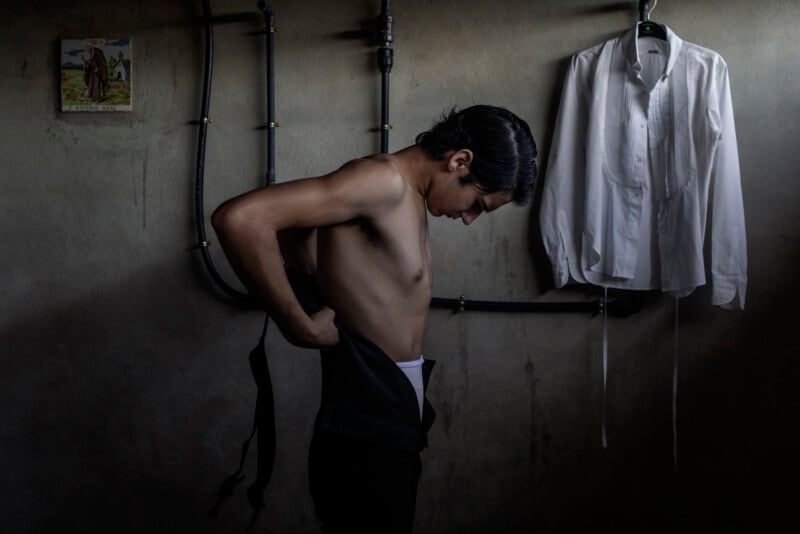 A shirtless young man stands in a dimly lit room, pulling on dark pants. A white dress shirt hangs on the wall beside him, and black pipes run along the wall in the background.