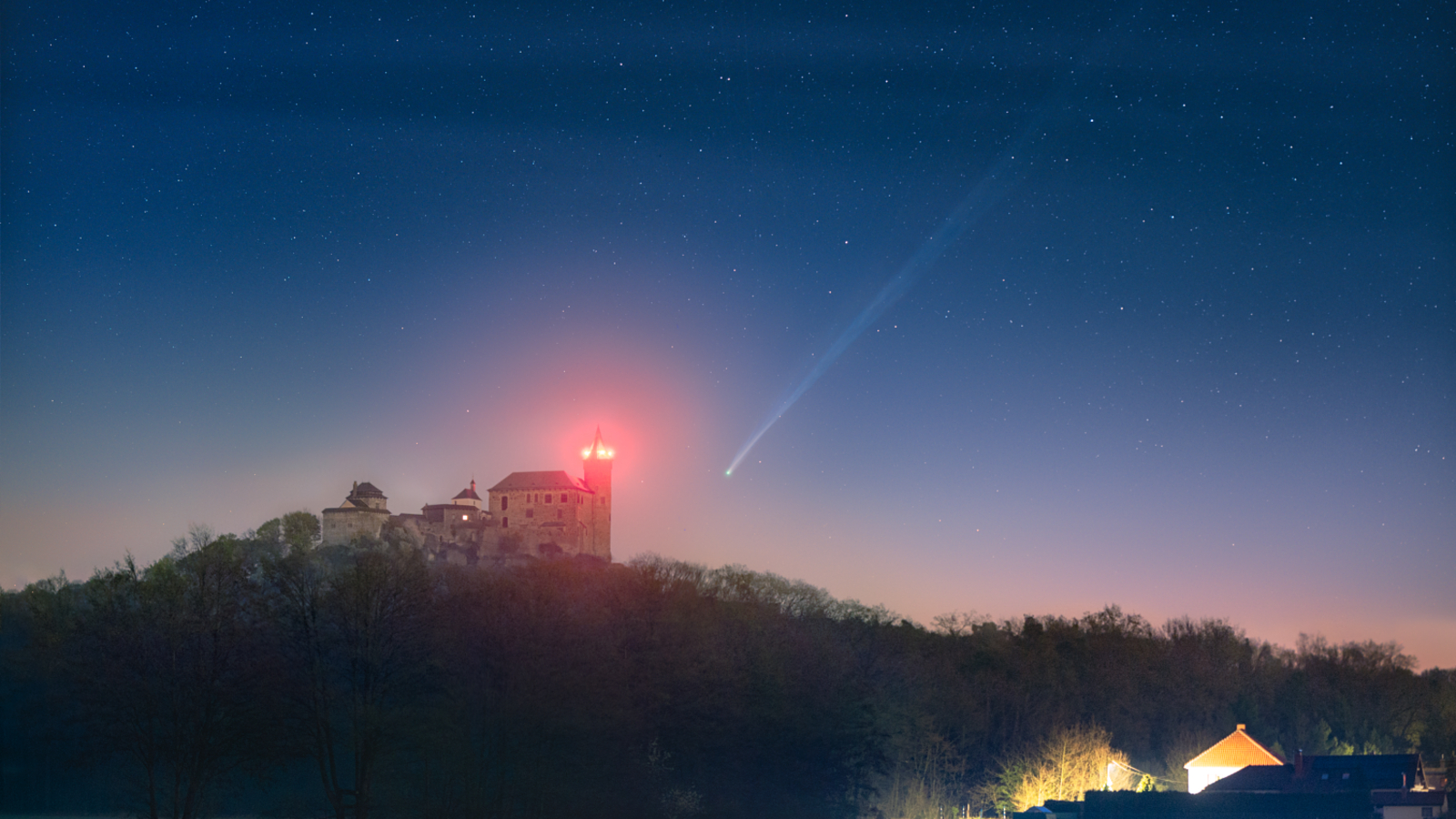 A photo of a long-tiled comet in the night sky. A medieval castle and house are illuminated in the foreground.