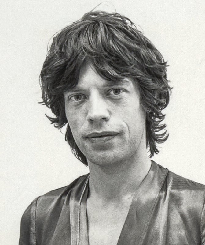 A person with shaggy, medium-length dark hair and light skin is wearing a shiny, open-collared shirt, looking directly at the camera with a neutral expression against a plain background.