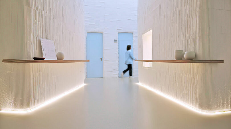 A person walks past two light blue doors in a minimalist, white hallway with floating shelves and soft floor lighting.