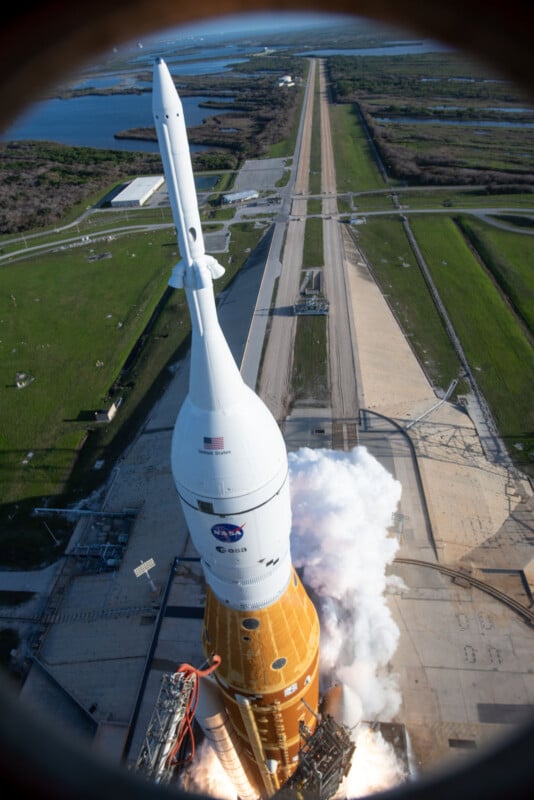 Aerial view of NASA’s Artemis I rocket launching from a launch pad, with white smoke billowing out and green fields and water visible in the background.