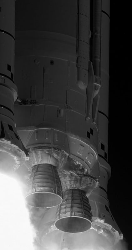 Close-up view of the bottom of a rocket at launch, showing three large engines firing bright flames and emitting smoke, with parts of the rocket body visible in detailed, high-contrast black and white.