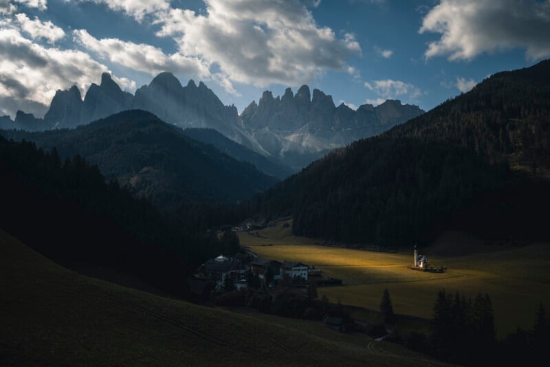 A sunlit church stands in a green valley surrounded by dark, forested hills, with dramatic, jagged mountains and scattered clouds in the background.