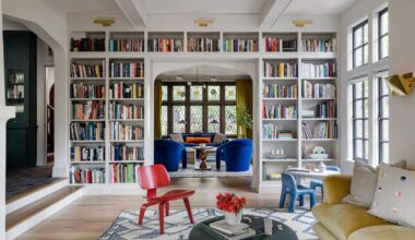 Living room with white painted walls, floor-to-ceiling bookcase with books, yellow velvet sofa, red chair on a rug, looking into another living room with blue armchairs and a blue sofa
