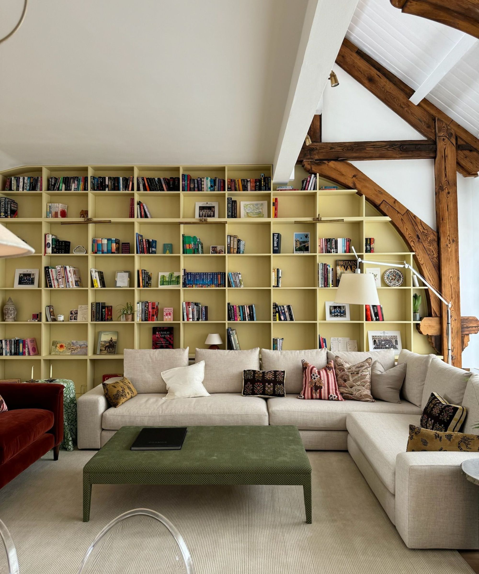 Living room with high beamed ceiling, cream sectional sofa, green ottoman, and a large built-in bookcase unit on the back wall painted lemon yellow and decorated with books, art and trinkets.