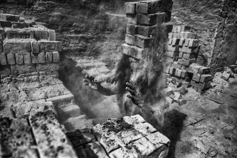 A person covered in dust, with only their hands and arms visible, emerges from a pile of bricks in a dim, dusty environment; bricks and debris surround them.