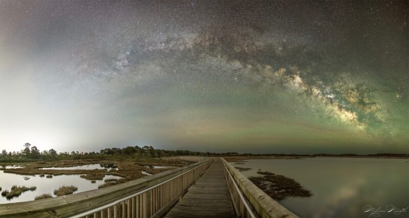 A boardwalk along water stretching away with the glowing, cloudy Milky Way arching overhead.