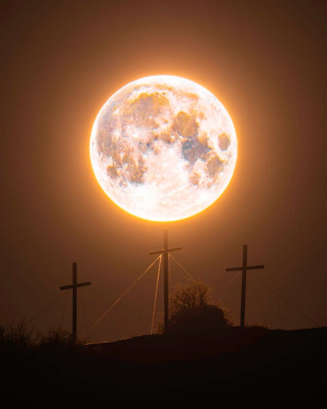 A bright full moon glows in the night sky, positioned directly above three silhouetted crosses on a hill, creating a dramatic and luminous scene with the moon appearing like a halo.