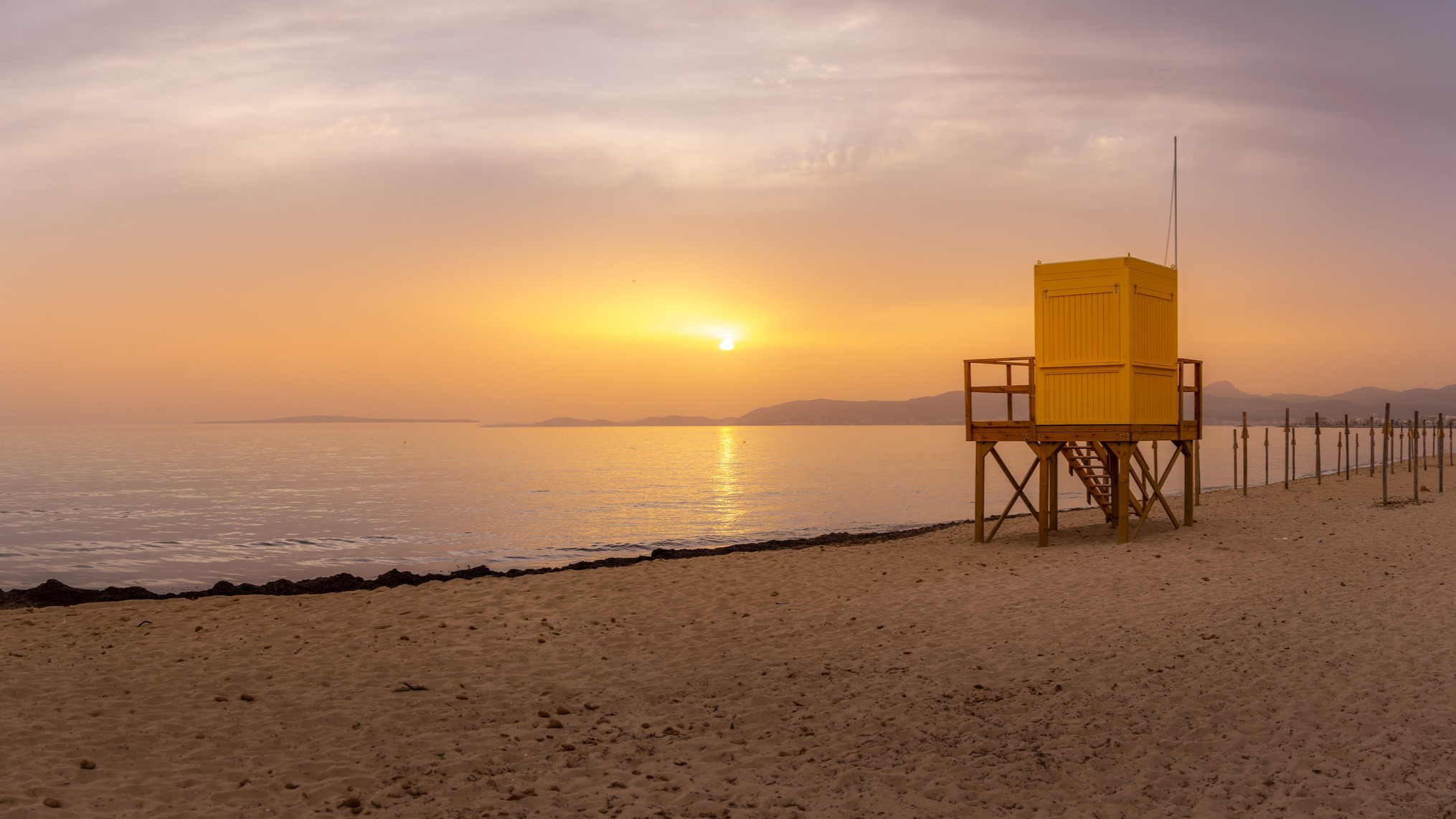 a lifeguard hut sits on a long sandy beach bathed in golden light from the setting sun.