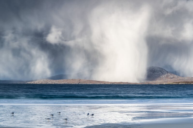 A dramatic coastal scene with dark storm clouds and a swirling column of snow over distant hills, turquoise waves in the foreground, and a few birds walking along the wet sandy beach.
