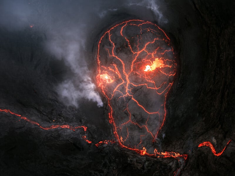 A glowing lava formation in a volcanic landscape, with cracks and fiery spots creating the eerie appearance of a skull-like face surrounded by dark smoke and cooled rock.