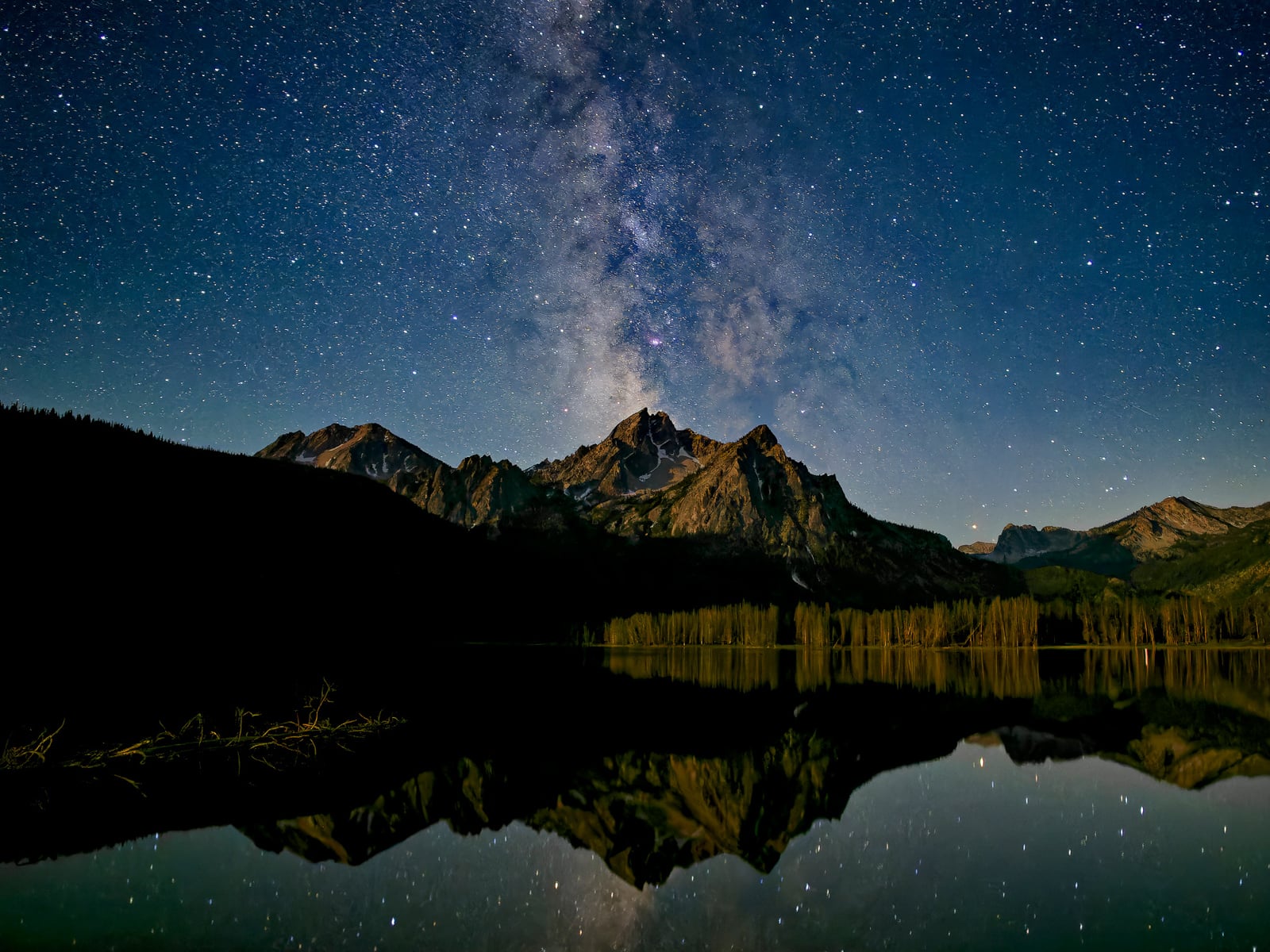 A star-filled night sky with the Milky Way visible above rugged mountain peaks, reflected in a calm lake below, surrounded by pine trees.