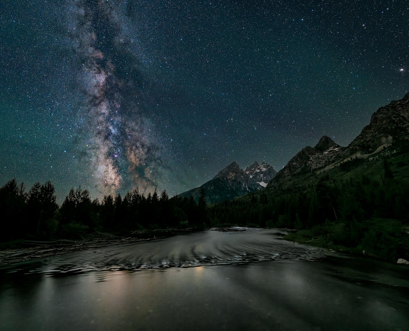 The Milky Way galaxy stretches brightly across a star-filled night sky above jagged mountain peaks and a calm reflective river bordered by dark pine trees.