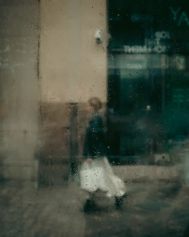 A blurred figure of a person in a white skirt and dark top is seen walking outside through a rain-soaked, foggy window, with water droplets on the glass. The background is urban and indistinct.