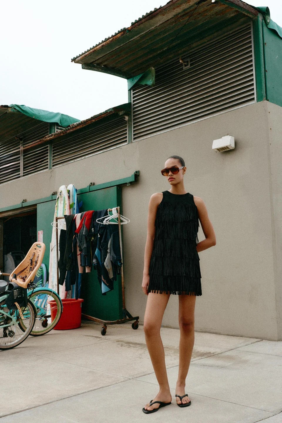 a person wearing a fringed black dress stands outside a building with surfboards and clothing hanging nearby