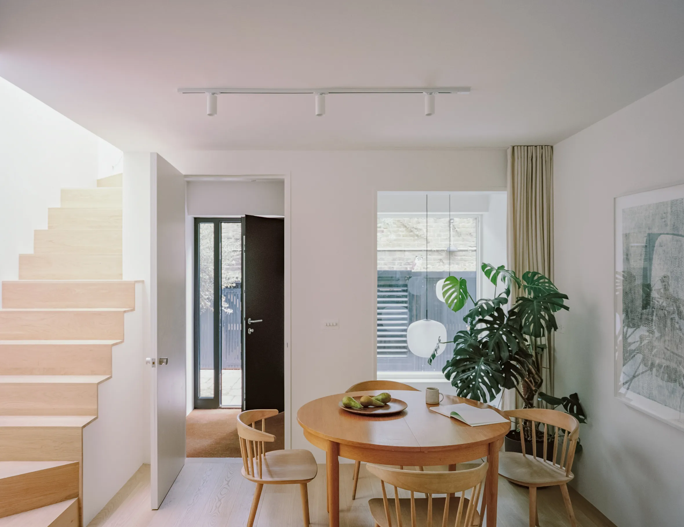 A dining room with a round wooden table and chairs, a large monstera plant, and a wooden staircase.