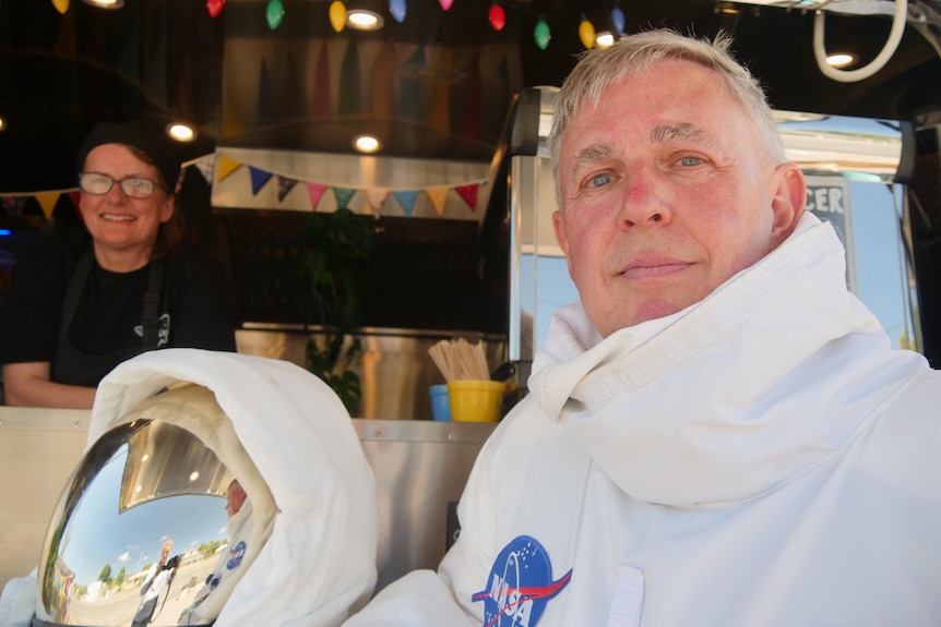 A man in a space suit stands next to a van done up to looks like a spaceship.