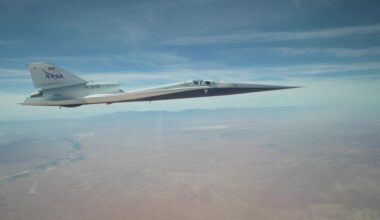 NASA’s X-59 quiet supersonic research aircraft flies above the Mojave Desert on a clear day. The aircraft is white with light gray, red, and blue accent colors. A NASA logo is visible on its tail, along with the number 859 above it. It appears to be flying level over the desert land-scape, and in the distance, you can see a mountain range on the horizon.
