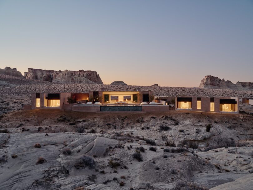 A modern, flat-roofed building with large glass windows is illuminated at dusk, set in a rocky desert landscape with mesas in the background.