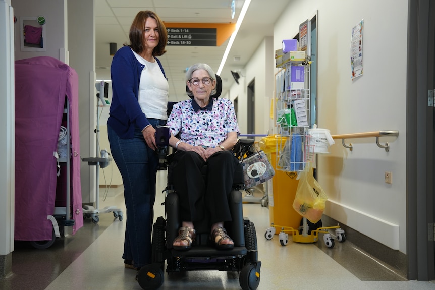 A woman with grey hair and glasses sits in a wheelchair, a woman with long brown hair standing next to her in a hospital.