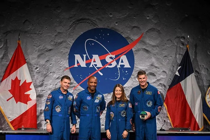 Four astronauts in blue jumpsuits stand in front of a NASA backdrop, with Canadian and American flags on either side