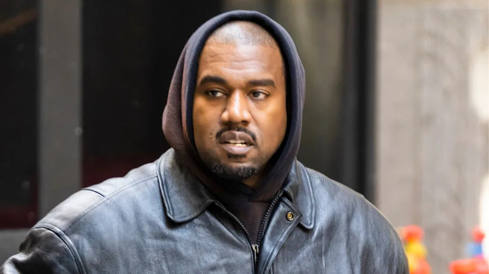 Kanye West in a black leather jacket and hoodie, walking outdoors.