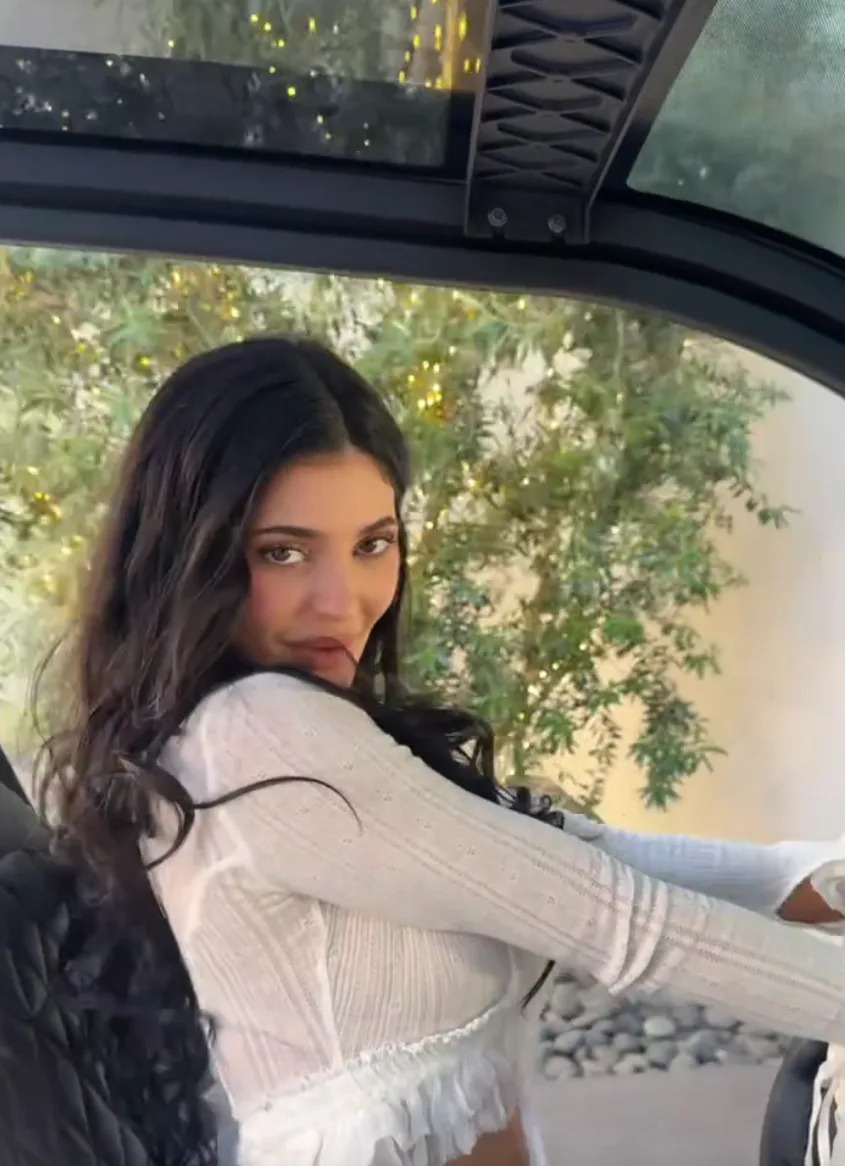Kylie Jenner with long, wavy hair wearing a stylish, fitted top, seated inside a vehicle, looking towards the camera with greenery visible in the background