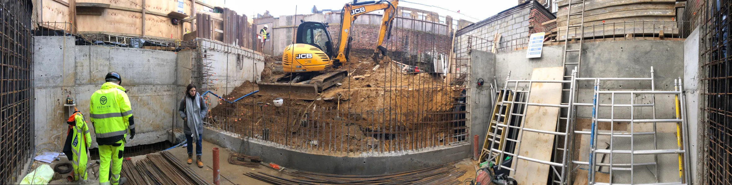 A construction site with an excavator in the center, and concrete walls, rebar, and construction workers around the perimeter.