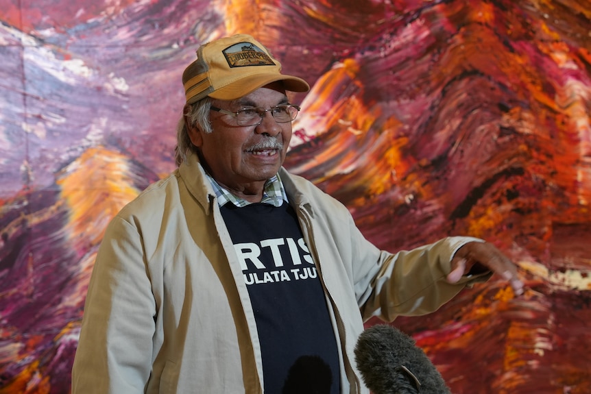 A man with a cap smiles in front of an artwork.