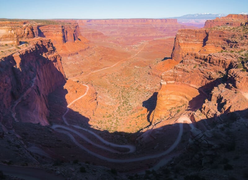 A winding dirt road curves through steep, red rock canyons under a clear blue sky, with rugged cliffs casting deep shadows over the landscape in a vast desert scene.