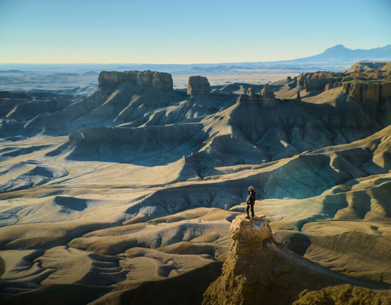 A person stands on a rocky outcrop overlooking a vast, rugged desert landscape with layered plateaus and distant mountains under a clear blue sky.