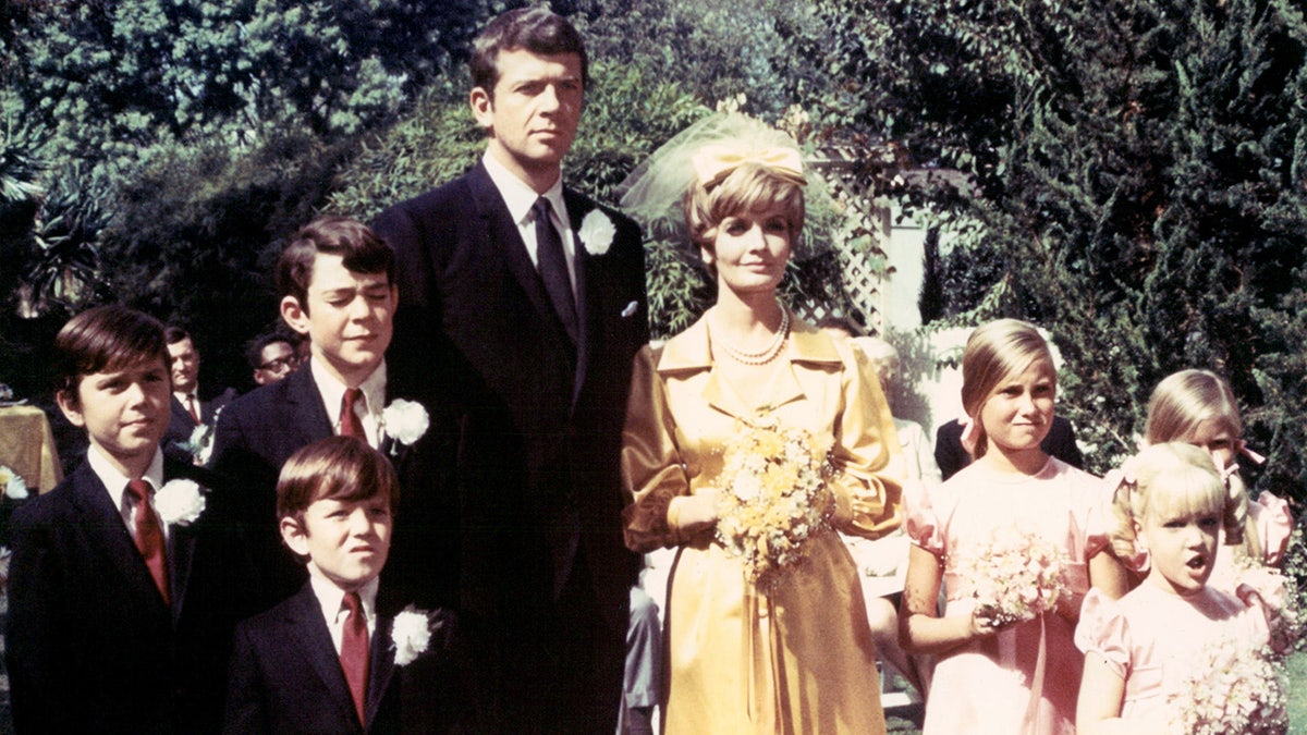 Barry Williams, Christopher Knight, Mike Lookinland, Robert Reed, Florence Henderson, Maureen McCormick, Eve Plumb, and Susan Olsen posing together at a wedding scene