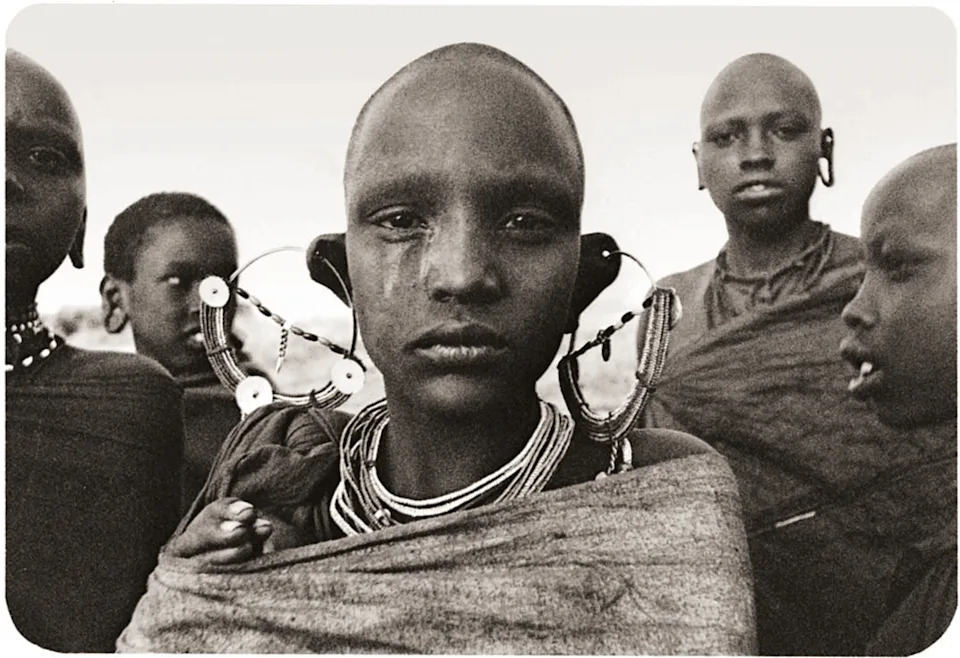 A black-and-white, close-up portrait of a young person with traditional stretched earlobes and ornate hoop earrings looking directly at the camera.