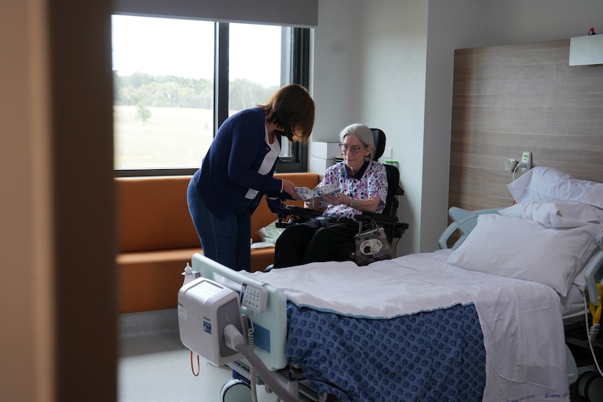 A woman with grey hair and glasses sits in a wheelchair, a woman with long brown hair leaning next to her in a hospital.