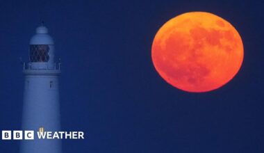 A red-orange full Moon hangs low in the night sky next to a lighthouse