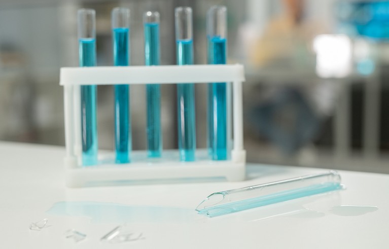 Five test tubes filled with blue liquid held in a white plastic rack, with one broken test tube lying on a white surface alongside scattered glass shards.