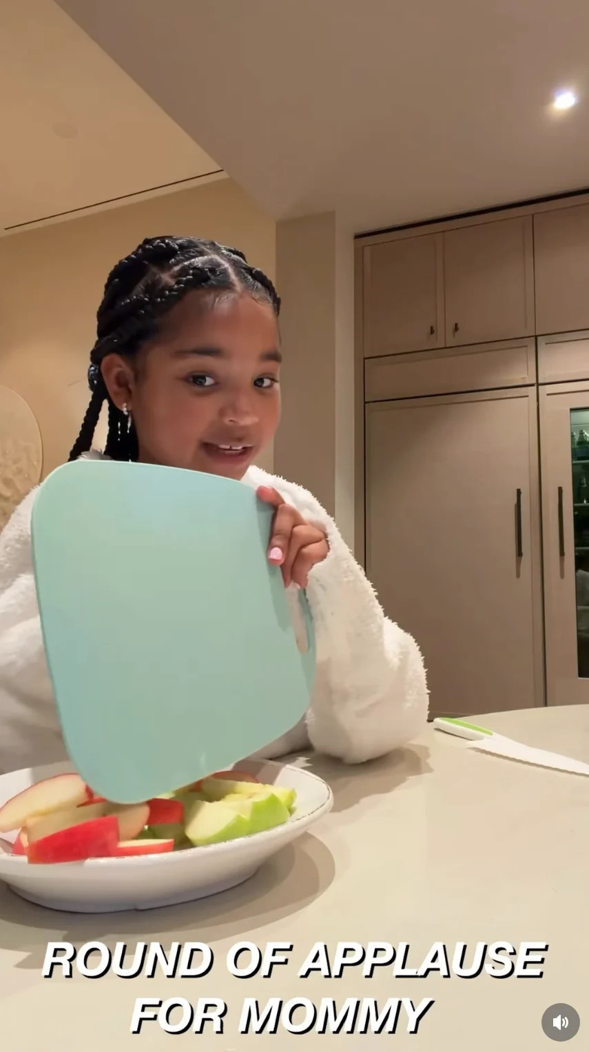 True smiling in a kitchen, holding a cutting board. Text: "Round of applause for Mommy"