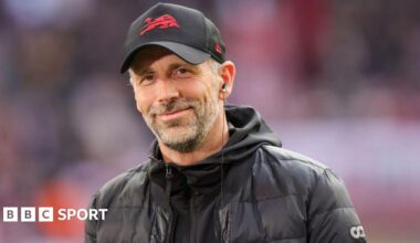 Marco Rose smiles and is wearing a baseball cap and a black coat