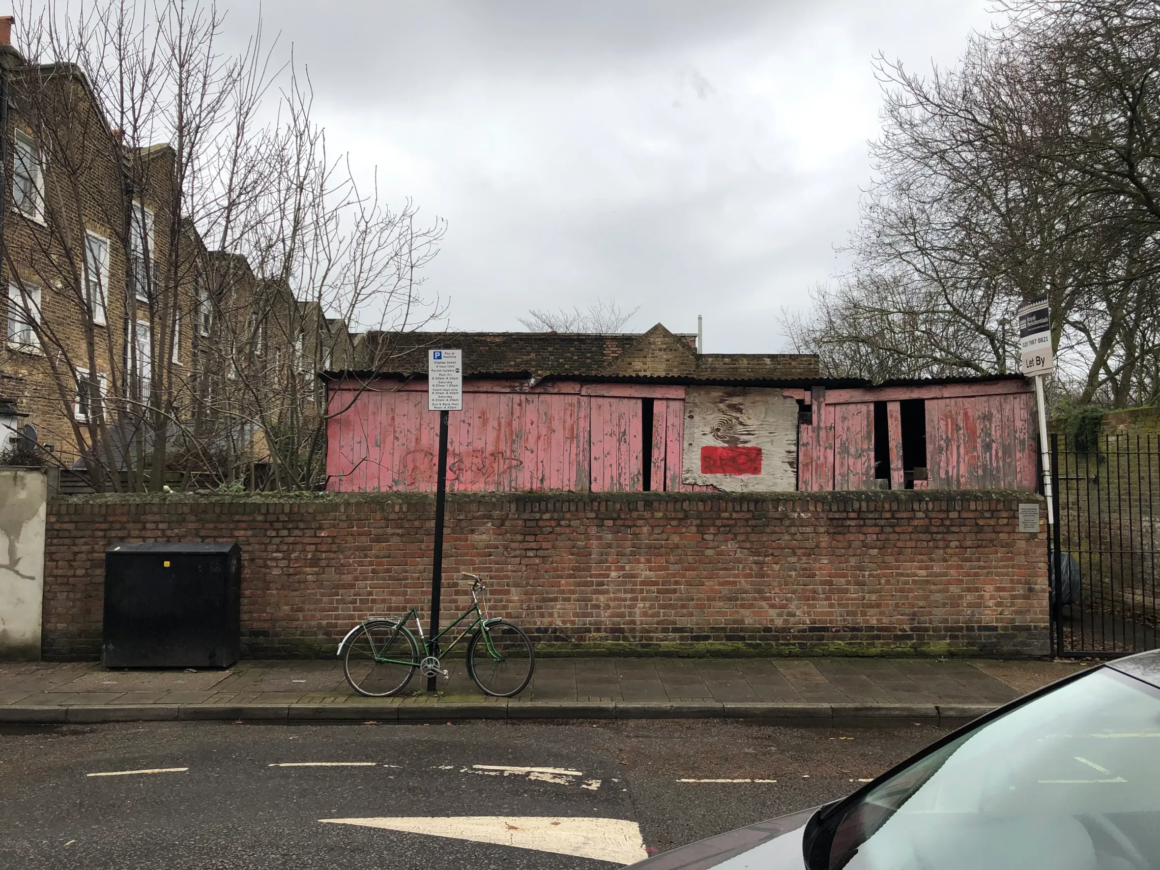 A pink shed with exposed wooden panels behind a brick wall, with a bicycle parked on the pavement.
